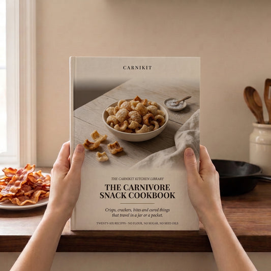 A person holds a hardcover cookbook titled "The Carnivore Snack Cookbook" by Carnikit in a warm kitchen setting. The book cover shows a bowl of nuts and crackers on a rustic table.