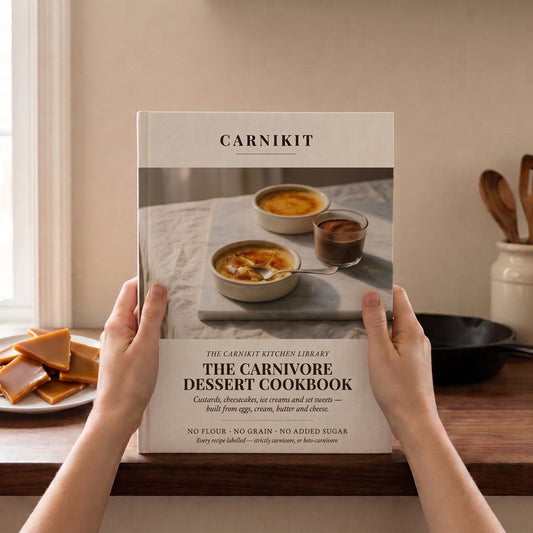 Two hands hold up a hardcover cookbook titled "Carnikit: The Carnivore Dessert Cookbook" against a warm-toned kitchen backdrop. The cover shows two crème brûlée-style custards in ramekins and a small cup of chocolate mousse on a marble board. Cover text reads "Custards, cheesecakes, ice creams and set sweets — built from eggs, cream, butter and cheese. No flour, no grain, no added sugar." A plate of caramel candies sits to the left; wooden spoons in a crock and a cast iron pan are visible on the right.