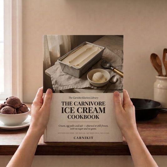 A person holds a hardcover cookbook titled "The Carnivore Ice Cream Cookbook" in front of a kitchen counter. The book cover shows a photo of an ice cream loaf tin and scoop, and the visible brand is Carnikit.