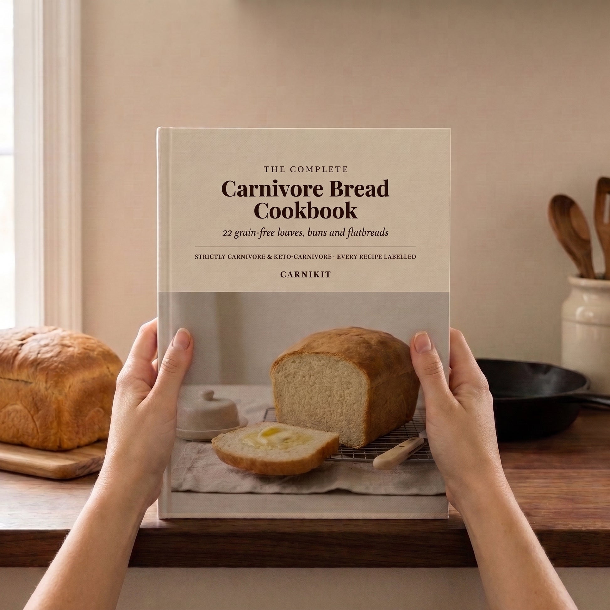 A person holds a hardcover cookbook titled "The Complete Carnivore Bread Cookbook" by Carnivite. The book cover shows a loaf of bread and a sliced piece on a kitchen counter, with a loaf of bread and a skillet visible in the background.