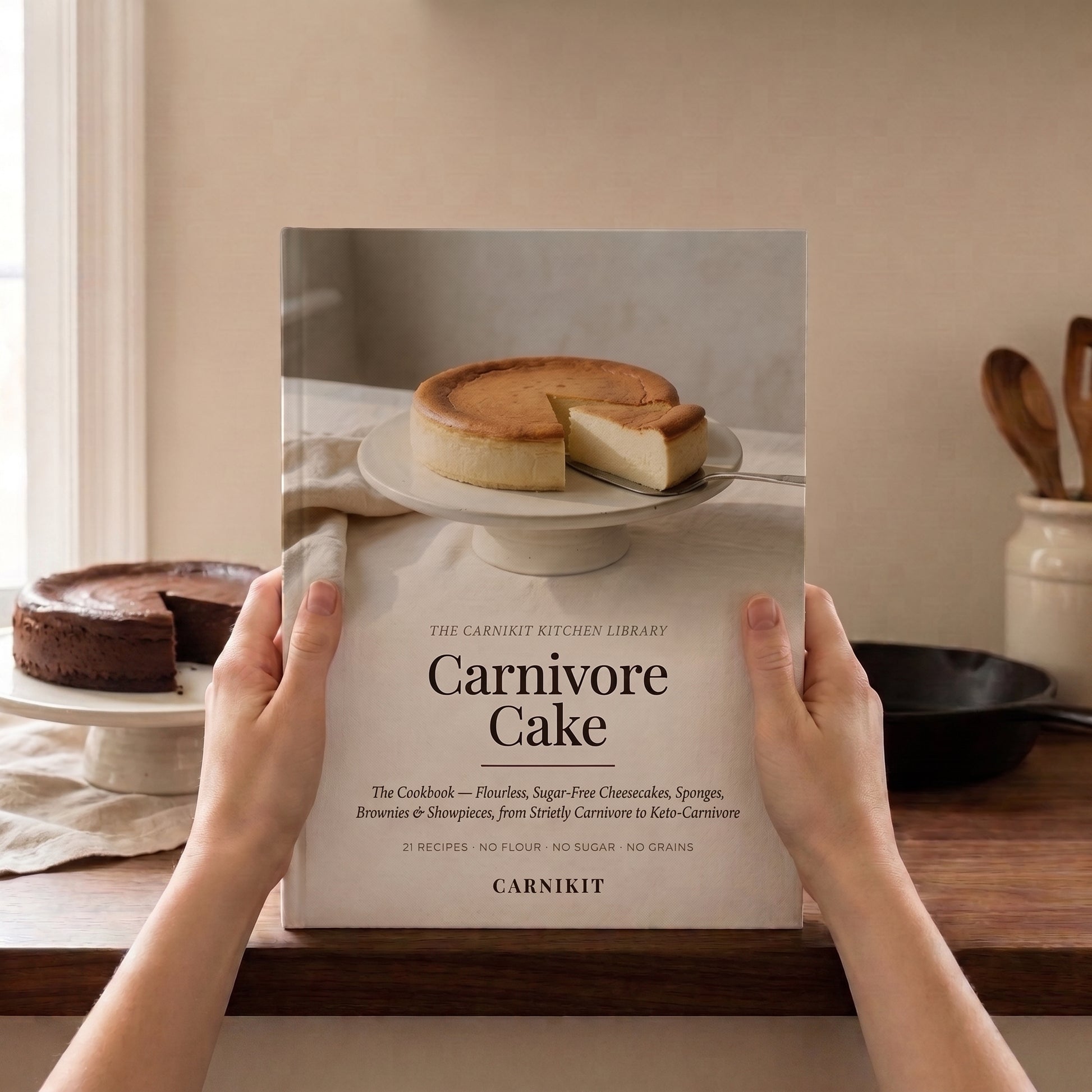 A person holds a cookbook titled "Carnivore Cake" in a kitchen setting. The book cover shows a cheesecake on a plate, and the visible text includes "The Carnivore Kitchen Library" and the brand "Carnikit" at the bottom.