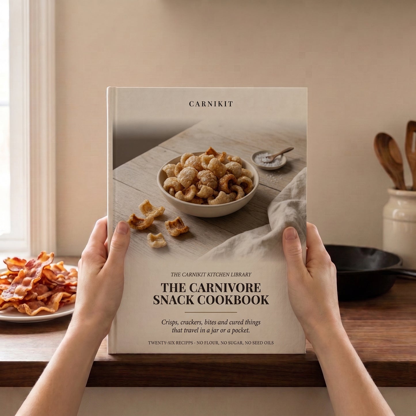 A person holds a hardcover cookbook titled "The Carnivore Snack Cookbook" by Carnikit in a warm kitchen setting. The book cover shows a bowl of nuts and crackers on a rustic table.