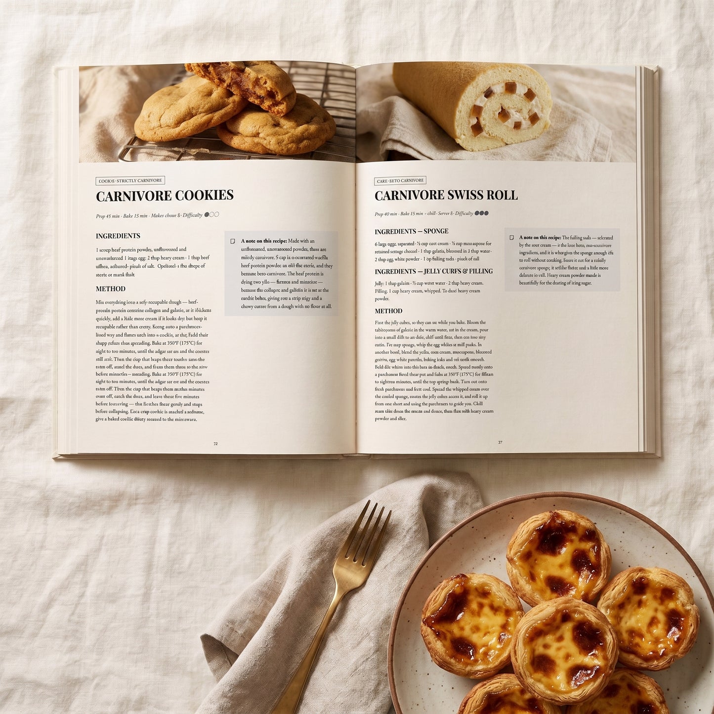 An open cookbook spread laid on a light fabric surface, showing recipe pages for carnivore cookies and a swiss roll, with a plate of baked tartlets and a gold fork nearby.