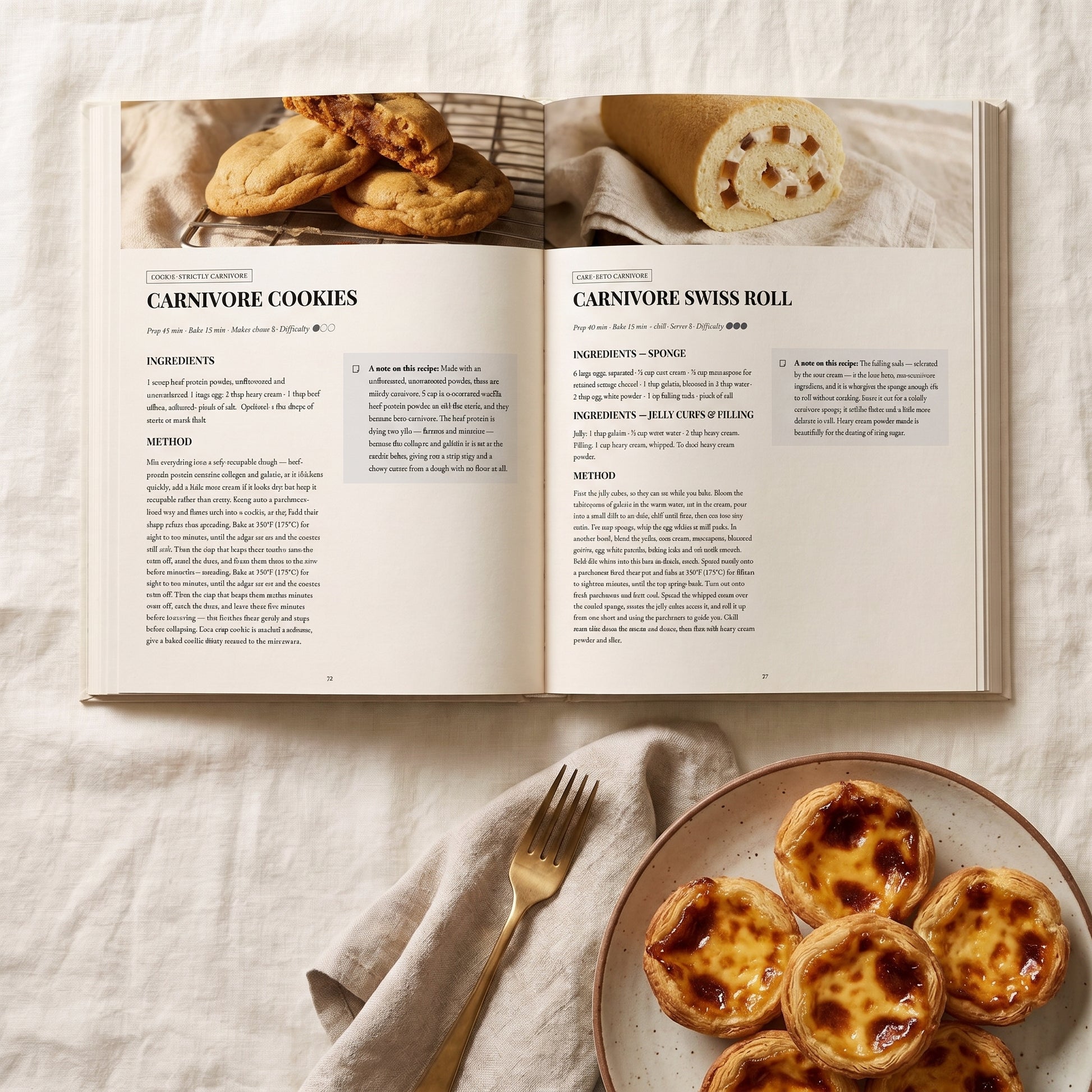 An open cookbook spread laid on a light fabric surface, showing recipe pages for carnivore cookies and a swiss roll, with a plate of baked tartlets and a gold fork nearby.