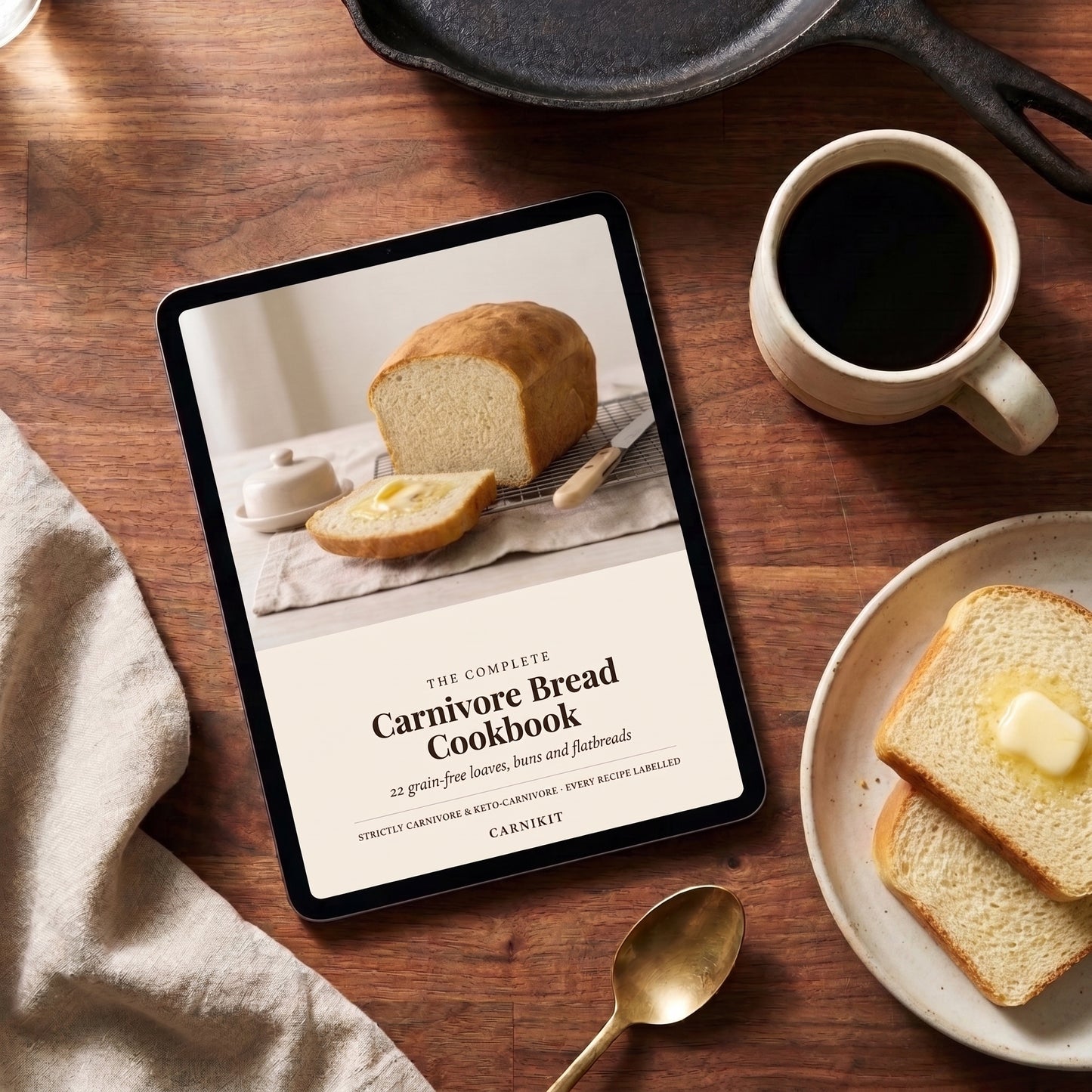 A tablet on a wooden table displaying a cookbook cover titled "The Complete Carnivore Bread Cookbook" by Carnivore. The scene is styled with a cup of black coffee, slices of bread with butter, a spoon, and a cast iron skillet.