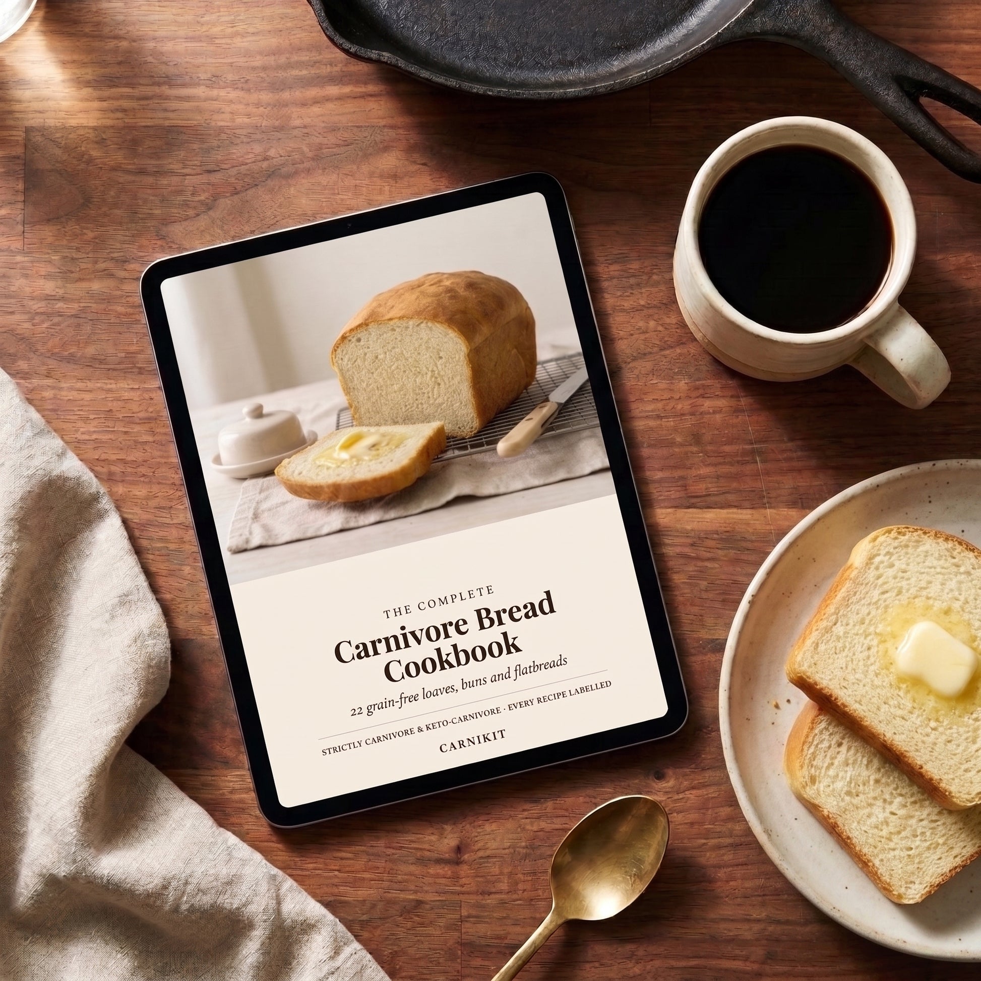 A tablet on a wooden table displaying a cookbook cover titled "The Complete Carnivore Bread Cookbook" by Carnivore. The scene is styled with a cup of black coffee, slices of bread with butter, a spoon, and a cast iron skillet.