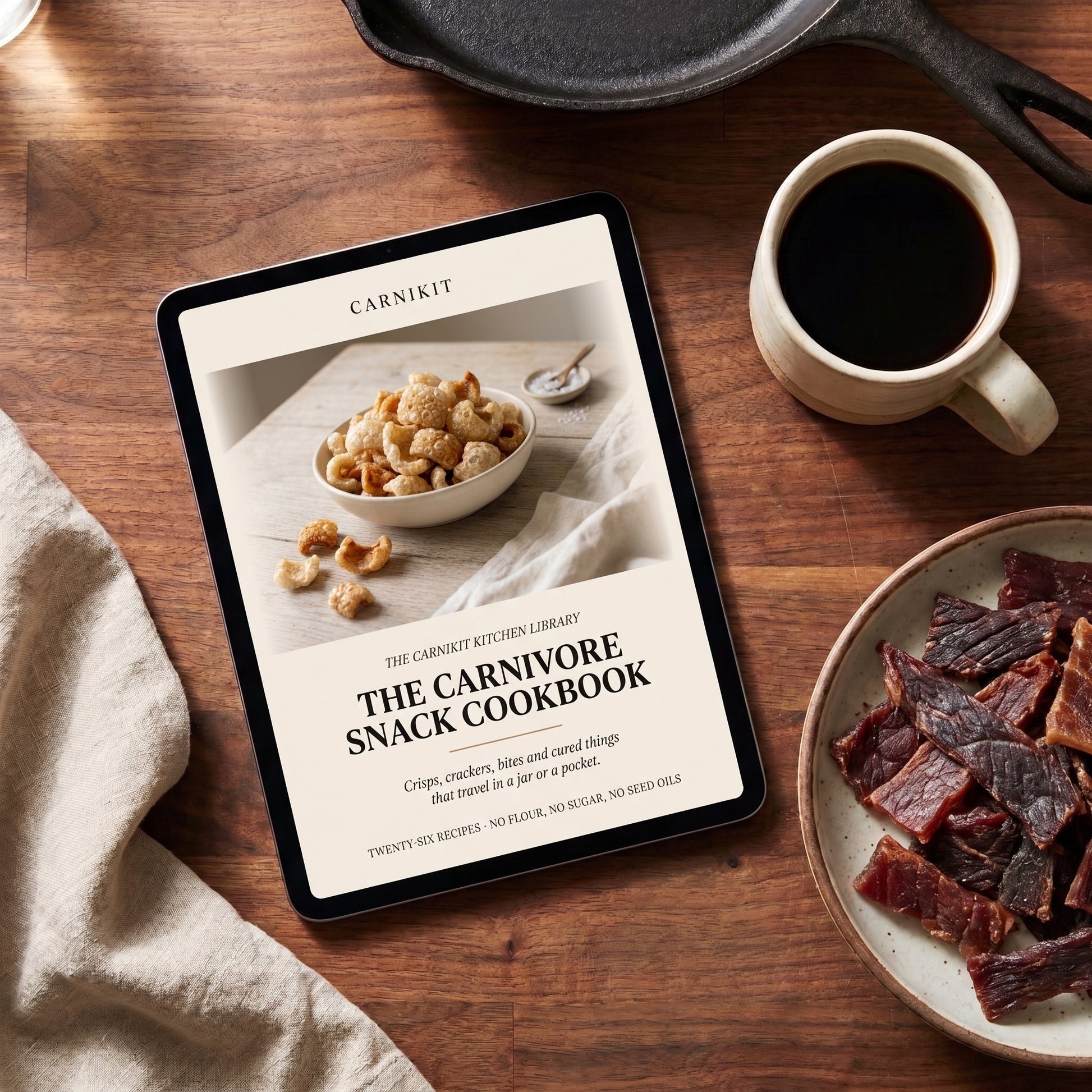 A tablet on a wooden table displaying the cover of 'The Carnivore Snack Cookbook' by Carnikit. The scene is styled with a cup of black coffee, a plate of beef jerky, a bowl of popcorn, and a cast iron skillet on a beige cloth.
