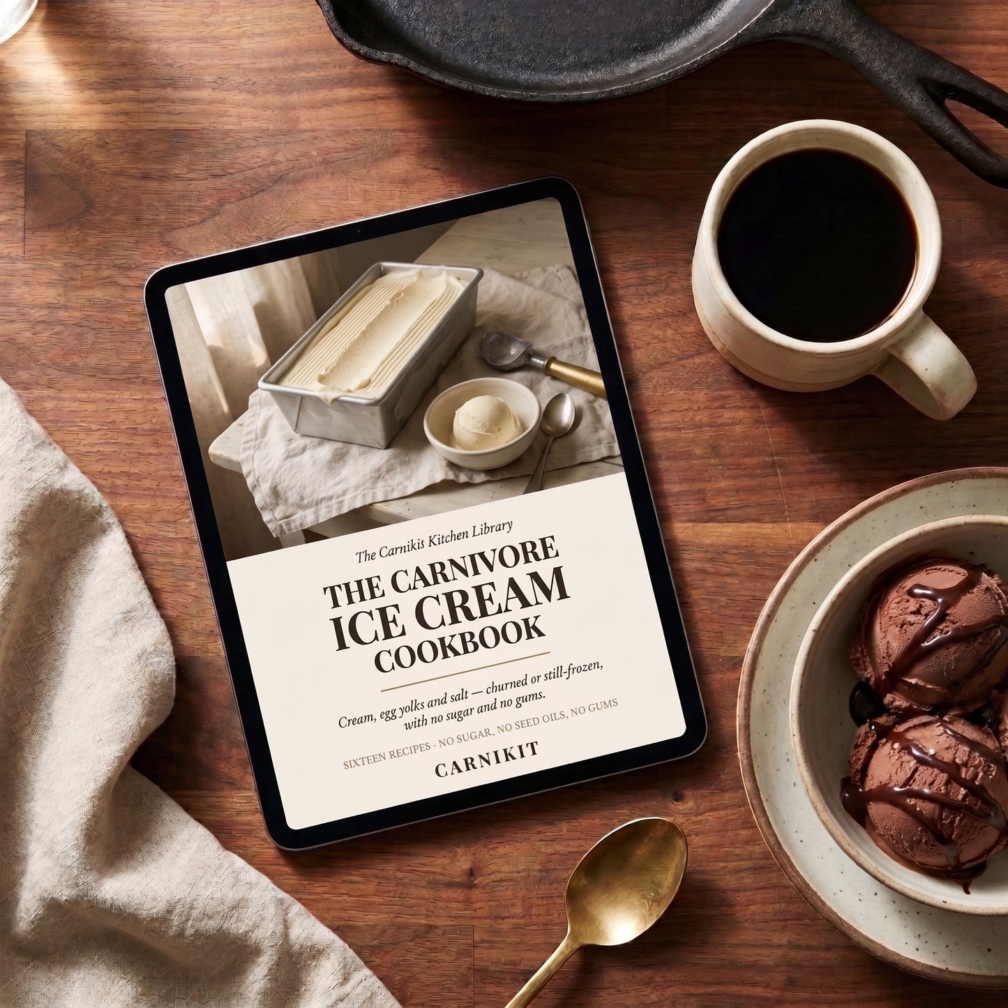 A tablet on a wooden table displaying the cover of 'The Carnivore Ice Cream Cookbook' by Carnikit. The scene is styled with a cup of black coffee, a bowl of chocolate ice cream, a gold spoon, and a beige cloth, with a cast iron skillet partially visible at the top.